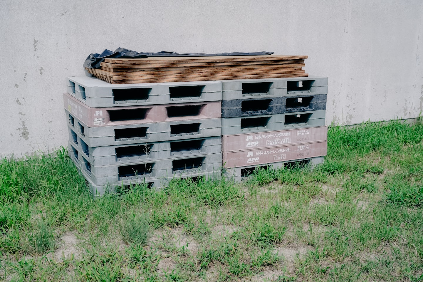 Plastic shipping pallets stacked on grass near a concrete wall.